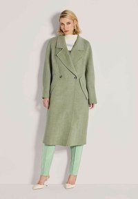 Vrouw in een lange groene dubbelzijdige jas, lichtgroene broek en crème hoge hakken staande tegen een effen beige achtergrond.