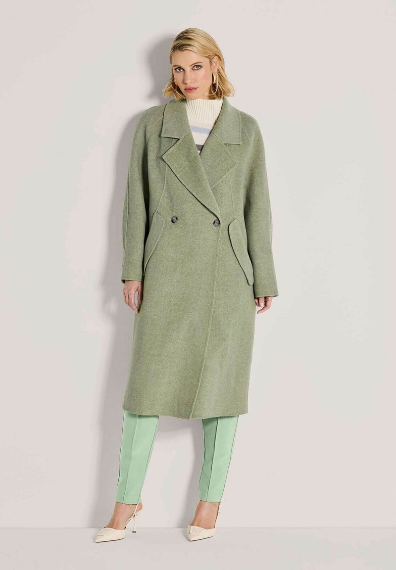 Vrouw in een lange groene dubbelzijdige jas, lichtgroene broek en crème hoge hakken staande tegen een effen beige achtergrond.
