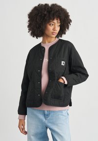 Giacca trapuntata nera con bottoni a pressione, dotata di due tasche frontali e una piccola toppa con il logo. Abbinata a una felpa rosa chiaro e jeans blu.