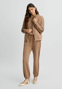 Bruine trainingsoutfit bestaande uit een zip-up jack met geribbelde manchetten en bijpassende broeken met elastische tailleband en zijstrepen. Compleet met platte schoenen met gouden accenten.