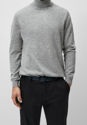 Mężczyzna w jasnoszarym golfie wpuszczonym w czarne spodnie z czarnym paskiem, z jedną ręką w kieszeni, na jednolitym tle.