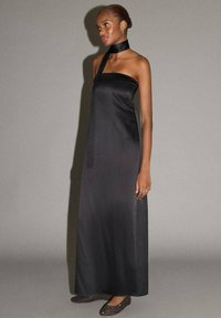 Robe maxi en satin noir avec un col licou et une texture lisse. Le mannequin porte des chaussures plates simples et un bracelet minimaliste.