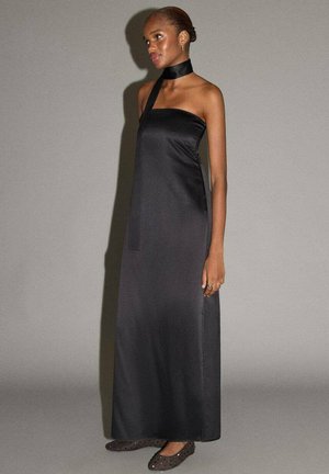 Abito lungo in raso nero con design a collo halter e texture liscia. La modella indossa scarpe semplici basse e un braccialetto minimalista.