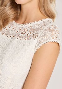 Weißes Spitzenoberteil mit floralen Motiven, gewellten Ausschnitt und kurzen Ärmeln. Das Material hat eine strukturierte, gewebte Optik.