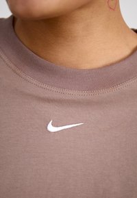 Brun kortärmad bomullströja med ribbat halsband och en vit Nike-logotyp broderad på bröstet. Slät textur.
