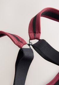 Gros plan de sangles rembourrées réglables couleur bordeaux et noir reliées par un clip en plastique noir, sur un fond clair uni.