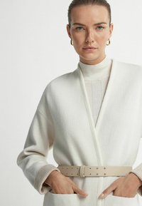Hvid uldblandingsfrakke med sjalskrave, sidelommer og et beige bælte. Indersiden har en lyse farvet turtleneck.