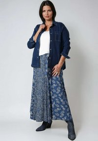 Woman wearing a dark blue denim shirt over white top, paired with a blue paisley and floral patterned long skirt and dark ankle boots.