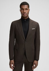 Brauner, maßgeschneiderter Anzug mit strukturierter Webart, einreihigem Design, Spitzrevers und weißem Einstecktuch. Getragen über einem schwarzen Reißverschluss-Top.