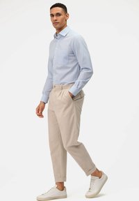 Lichtblauwe, lange mouwen overhemd met een borstzak, gecombineerd met beige geplooide broeken en witte sneakers, op een figuur tegen een effen achtergrond.