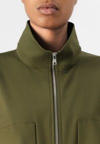 Chaqueta verde oliva con un cuello alto, cierre de cremallera y dos bolsillos frontales, hecha de una tela suave y texturizada.