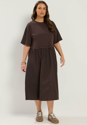 Mujer con pelo largo lleva un vestido midi suelto de color marrón oscuro y zapatillas beige, de pie frente a un fondo liso y claro.