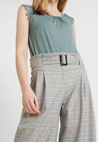 Grön ärmlös topp med spetsdetaljer; vida rutiga byxor i beige, blå och brun med bälte i midjan och pressesvik fram.