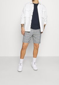 Biała koszula na guziki z dwoma kieszeniami na piersi, granatowy t-shirt, szare szorty w kraty, białe trampki i białe skarpetki. Casualowy letni strój.