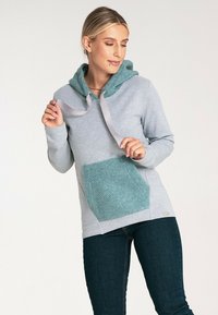 Szary sweter z kapturem w kolorze teal i podszewką z polaru oraz kieszenią. Posiada satynowe sznurki i miękką teksturę. Noszony z ciemnymi dżinsami.