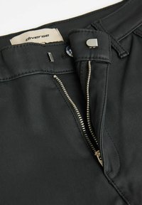 Czarne spodnie wykonane z gładkiego materiału, z zamkiem błyskawicznym i metalowymi elementami wykończenia, z metką z napisem "diverse" w pobliżu pasa.