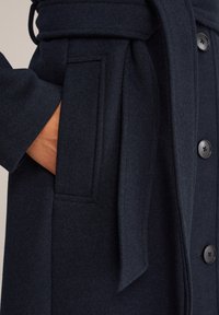 Mörk ullkapp med bälte i midjan. Har en ficka med patch, stora svarta knappar och en mjuk, texturerad yta.