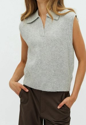 Ärmelloses graues, geripptes Stricktop mit Kragen, getragen zu braunen Hosen, Hände in den Taschen, Modell von Hals bis Hüfte gezeigt.