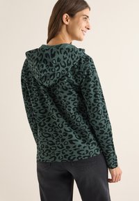 Felpa verde con stampa leopardata nera, dotata di cappuccio con lacci e vestibilità comoda. Tessuto morbido; indossata con jeans scuri.