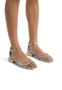Gouden platte sandalen met banden hebben een geweven ontwerp, open teen, enkelband met gespsluiting en een lage blokhak. Glad, metallic afwerking.