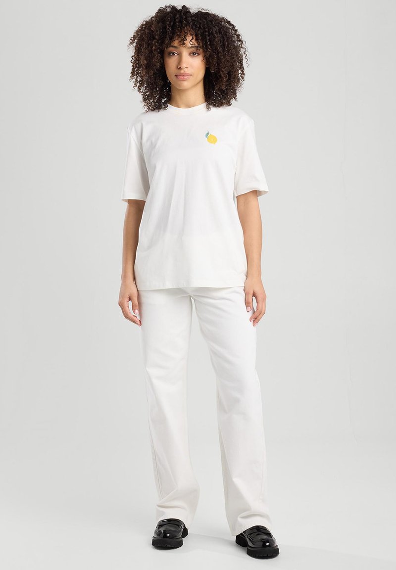 White short-sleeve t-shirt featuring a small lemon graphic, paired with white flared pants and black shoes. Smooth fabric texture.