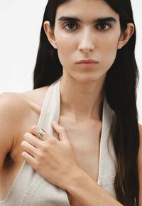 Anillo de plata y oro en capas, llevado en una mano de tono de piel claro, contra un fondo neutro. La persona que lo lleva tiene el cabello liso y oscuro.