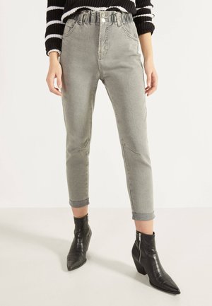 Femme portant un jean gris taille haute avec des revers retroussés et des bottines noires à bout pointu avec des fermetures éclair latérales.