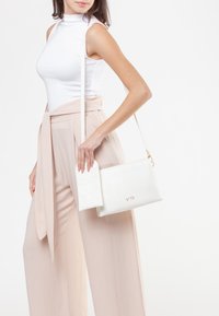 Borsa a tracolla bianca in pelle con una patta strutturata e dettagli in oro. Il modello indossa un top bianco e pantaloni rosa chiaro, mettendo in risalto il design della borsa.