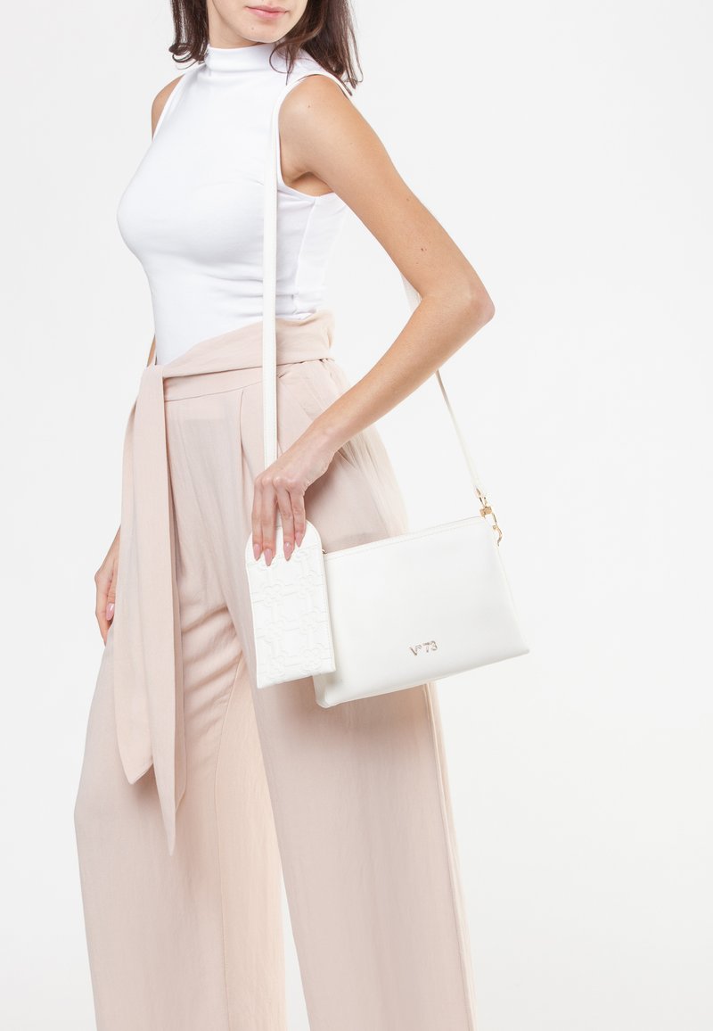 Borsa a tracolla bianca in pelle con una patta strutturata e dettagli in oro. Il modello indossa un top bianco e pantaloni rosa chiaro, mettendo in risalto il design della borsa.