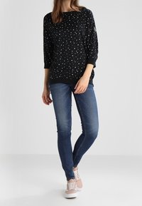 Kobieta ubrana w czarną wzorzystą bluzkę z długim rękawem, niebieskie wąskie dżinsy i jasnoróżowe trampki, stojąca ze skrzyżowanymi nogami.