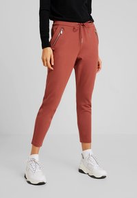 Joggingbroeken in roestkleur met een taps toelopende pasvorm, twee zijzakken met rits en een elastische tailleband met trekkoord. Gecombineerd met witte sneakers.