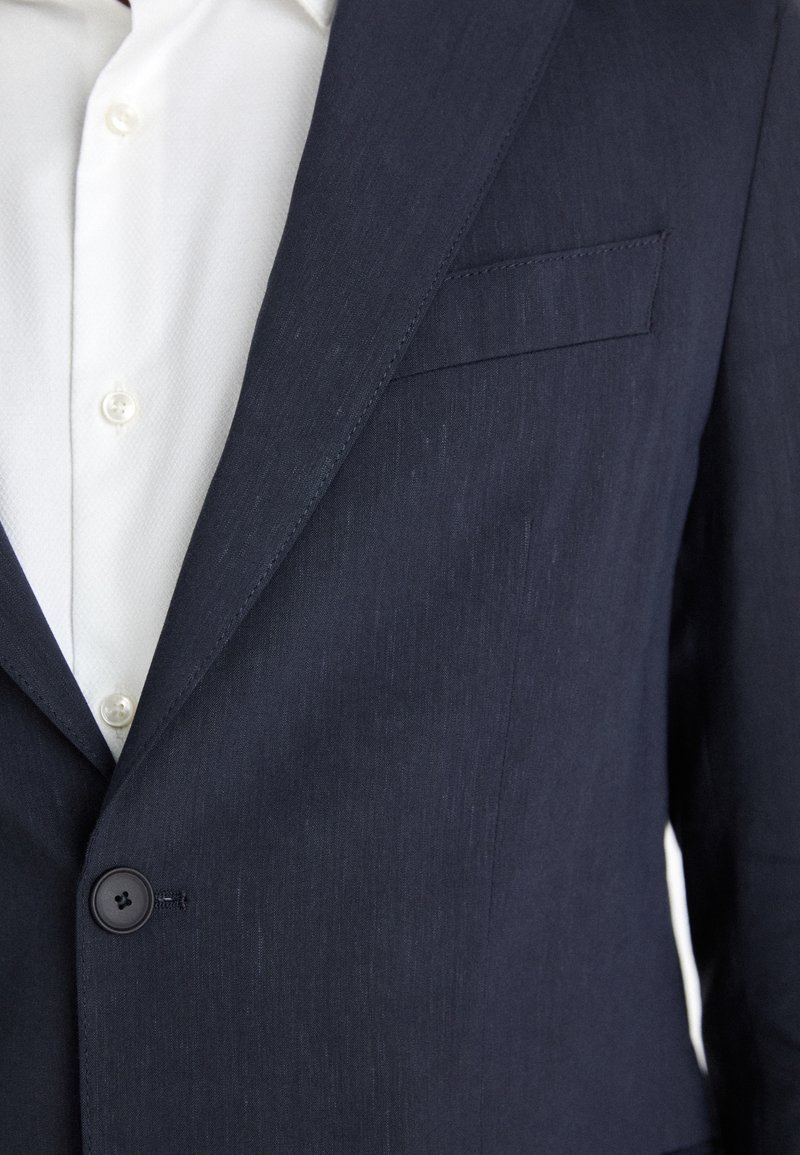 Veste de costume bleu marine avec une finition texturée, fermeture à un bouton et poche poitrine. Portée sur une chemise blanche avec des boutons visibles.