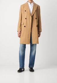 Cappotto doppiopetto in lana color cammello con revers a lancia, due tasche frontali e bottoni neri. Abbinato a jeans di jeans blu e scarpe da ginnastica nere.