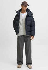 Marineblå pufferjakke, grå strik cardigan, hvid skjorte, grå brede bukser og beige slip-on sko; glatte teksturer og afslappet pasform.