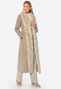 Beige lange jas met een luchtige nepbontkraag, recht design, zijzakken en een subtiele textuur. Draagt over lichtbeige broeken.