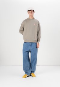 Grå hettegenser med en liten rød logo, kombinert med løse blå jeans og gule joggesko. I tillegg har den en lysfarget caps.