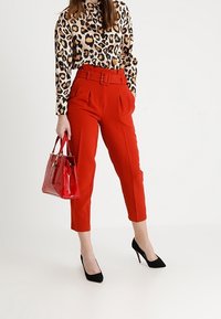 Pantaloni a vita alta rossi con pieghe, abbinati a una blusa con stampa leopardata e décolleté nere. Presenta anche una borsa rossa lucida.