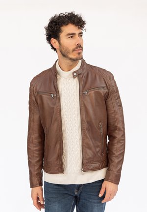 Hombre con cabello rizado que lleva una chaqueta de cuero marrón, suéter crema de punto trenzado y vaqueros azules, mirando hacia un lado.