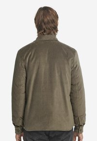 Widok z tyłu osoby ubranej w oliwkową koszulę z korduroyu z guzikami na mankietach oraz czarne dżinsy na białym tle.