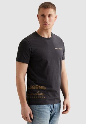 Młody mężczyzna w czarnej koszulce PME Legend i niebieskich dżinsach, patrzący na bok na jednolitym, jasnoszarym tle.