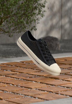 Baskets basses noires avec semelle blanche et bout renforcé blanc, montrées en l'air au-dessus d'une surface extérieure en bois et gravier à motif.