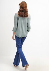 Blusa verde chiaro con tessuto testurizzato, maniche a tre quarti e vestibilità comoda, abbinata a jeans svasati blu scuro e sandali.
