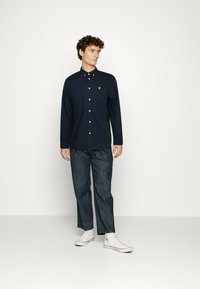Camisa de manga larga azul marino con un pequeño logo amarillo en el bolsillo del pecho, combinada con pantalones de mezclilla oscuros y zapatillas deportivas blancas.