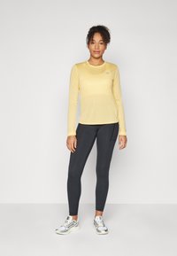 Langermet, lysegul treningstopp med rund hals; kombinert med mørke tights med sidelommer og grå joggesko.