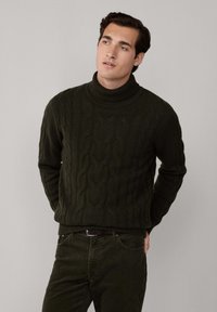 Man i mörkgrön kabelstickad polotröja och matchande byxor, stående med händerna bakom ryggen mot en enfärgad bakgrund.