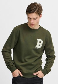 Oliven grøn sweatshirt med rund udskæring, ribstrikkede manchetter og et hvidt blokbogstav "B" broderet på venstre bryst. Blød stoftekstur.