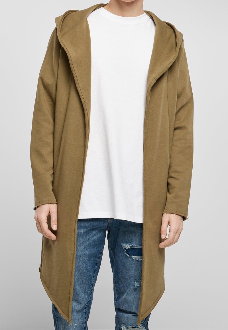 Cárdigan largo marrón con capucha, hecho de un tejido suave, que presenta un diseño abierto y un ajuste relajado, combinado con una camiseta blanca y jeans desgastados.