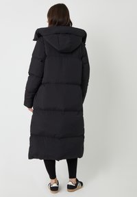 Zwart lange pufferjas met een capuchon, voorzien van een gewatteerd ontwerp en een oversized pasvorm, gedragen over zwarte leggings en casual schoenen.
