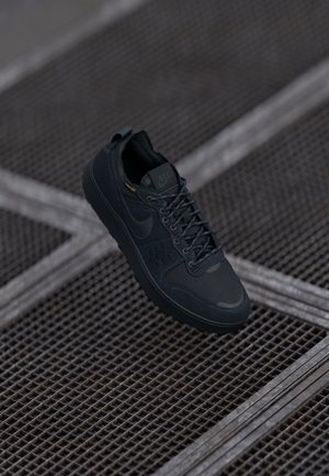 Czarny sneaker Nike z teksturowaną podeszwą, cholewką z siateczki i skóry oraz sznurowadłami, umieszczony na tle metalowej kratownicy podłogowej.
