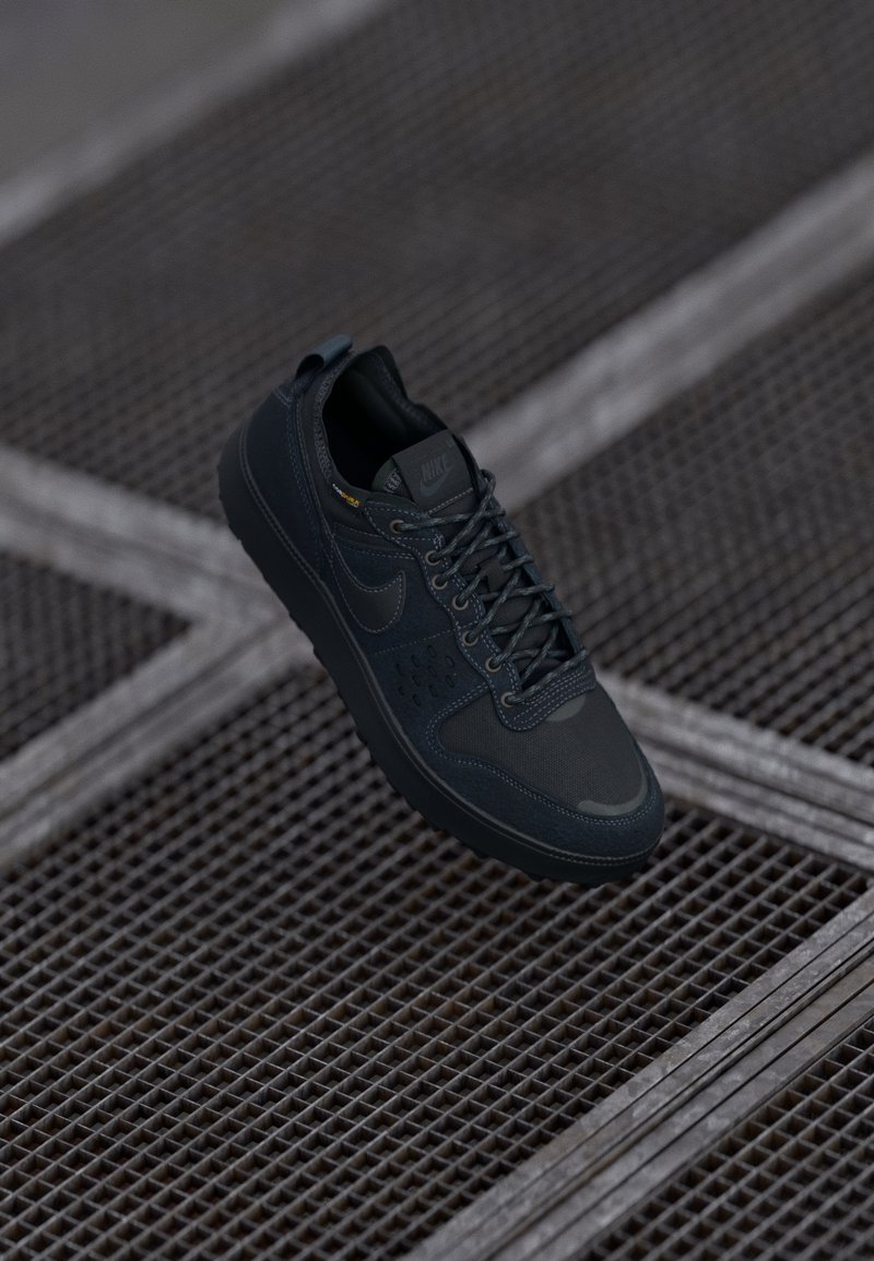 Baskets Nike noires avec semelle texturée, dessus en mesh et cuir et lacets, positionnées au-dessus d’un sol en grille métallique.